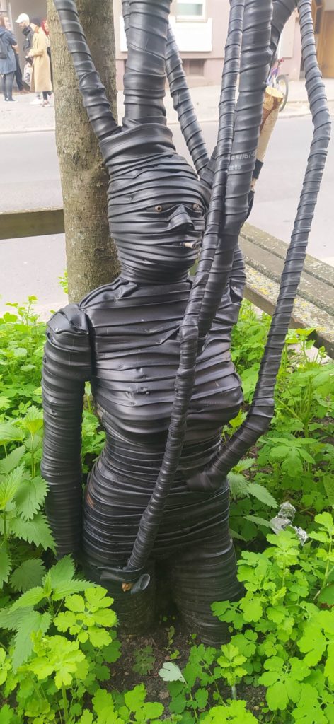 Recyled Jewellery: A creative sculpture made from black, corrugated flexible pipes, shaped into a humanoid form with intertwined arms and a coiled torso, surrounded by green foliage on a city street. This artistic piece incorporates @ Reblack Shop